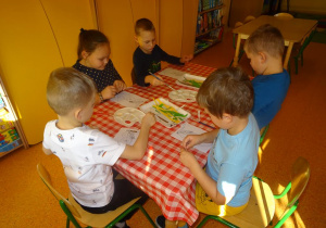 Piątka dzieci siedzi przy stoliku, wykleja wydzieranymi kawałkami papieru sylwetę Syrenki Warszawskiej.